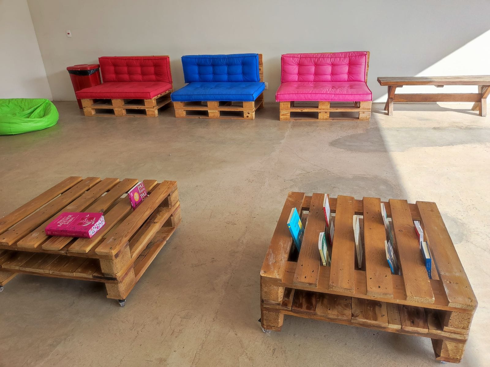 Sala de descanso simples com sofás de pallet coloridos, banco de madeira e mesas de centro de pallet com rodízios